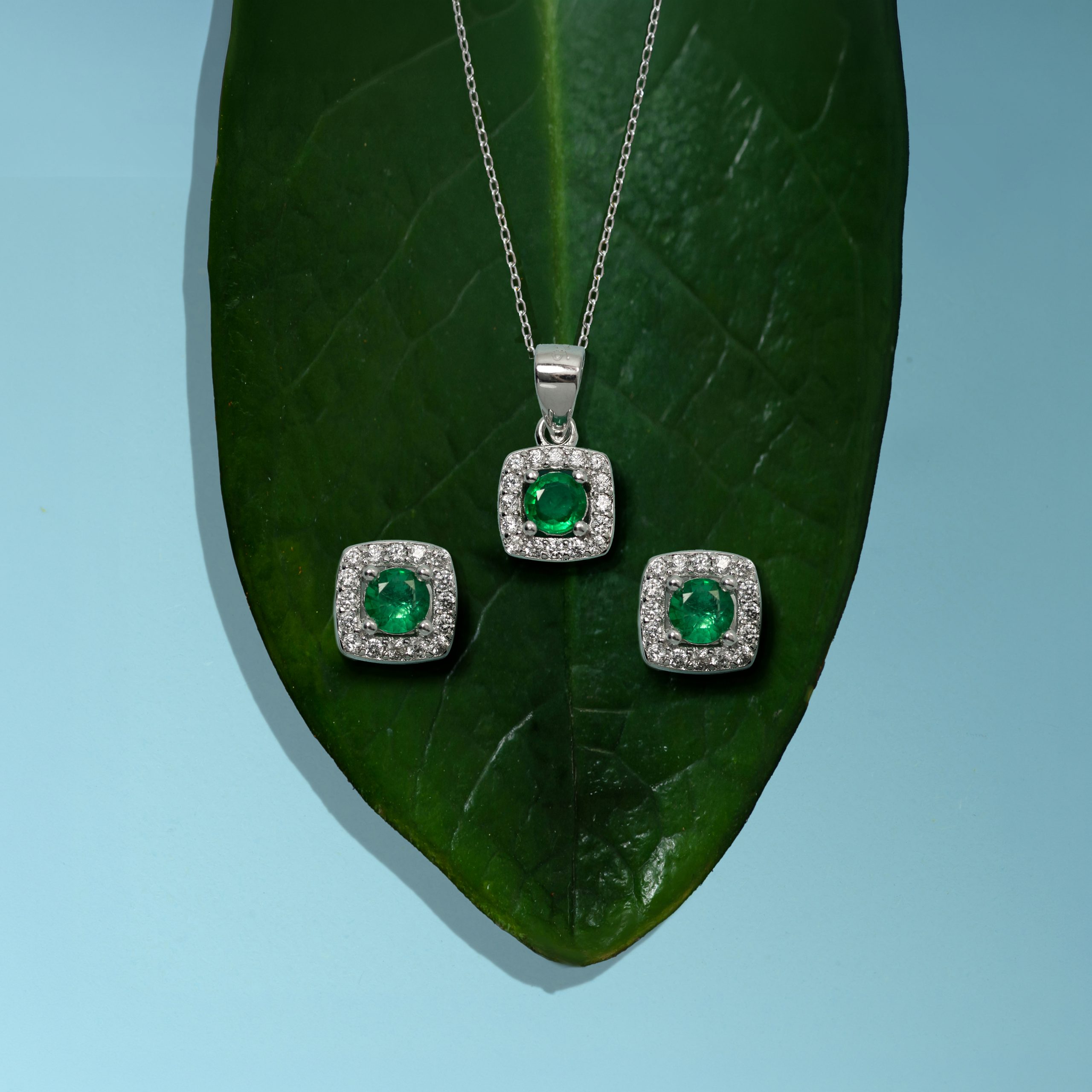_NZR1377 Silver jewelry set with green emerald stones and diamond accents displayed on a large green leaf on a blue background.