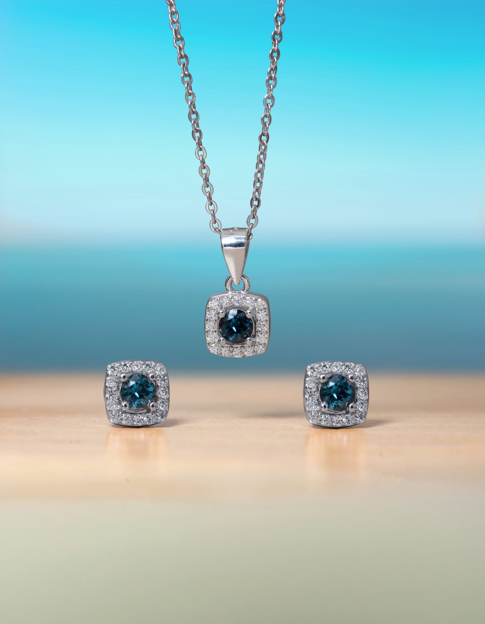 Half Set 2 Silver necklace with a square pendant showcasing a blue gemstone surrounded by small diamonds, against a blue gradient backdrop.