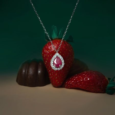 Close-up of a red strawberry wrapped with a silver chain necklace featuring a pink teardrop pendant encrusted with small stones.