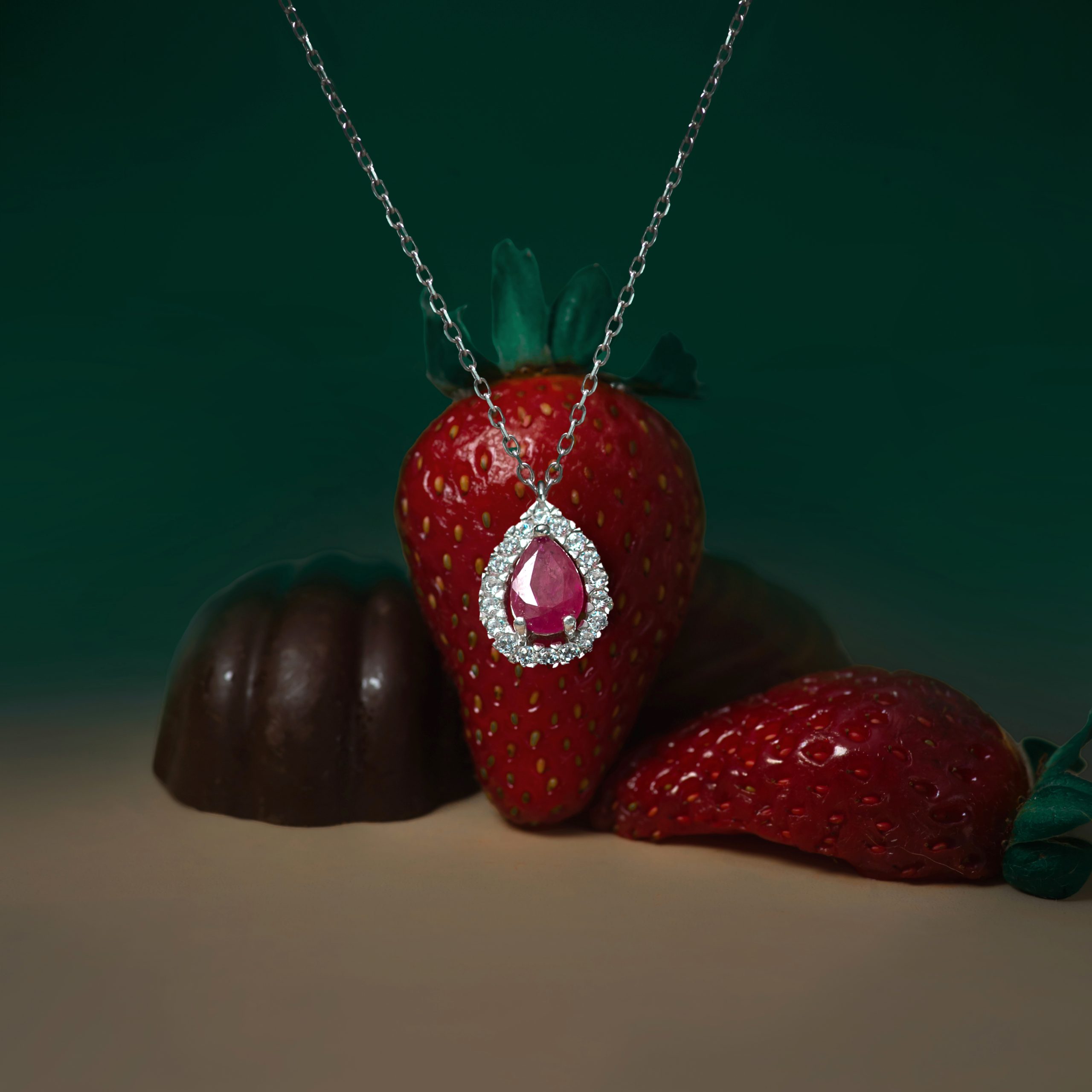 _NZR9932v Close-up of a red strawberry wrapped with a silver chain necklace featuring a pink teardrop pendant encrusted with small stones.