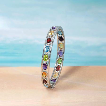 Curved silver bracelet with rainbow-colored gemstones standing on a sandy beach with a blue ocean backdrop.