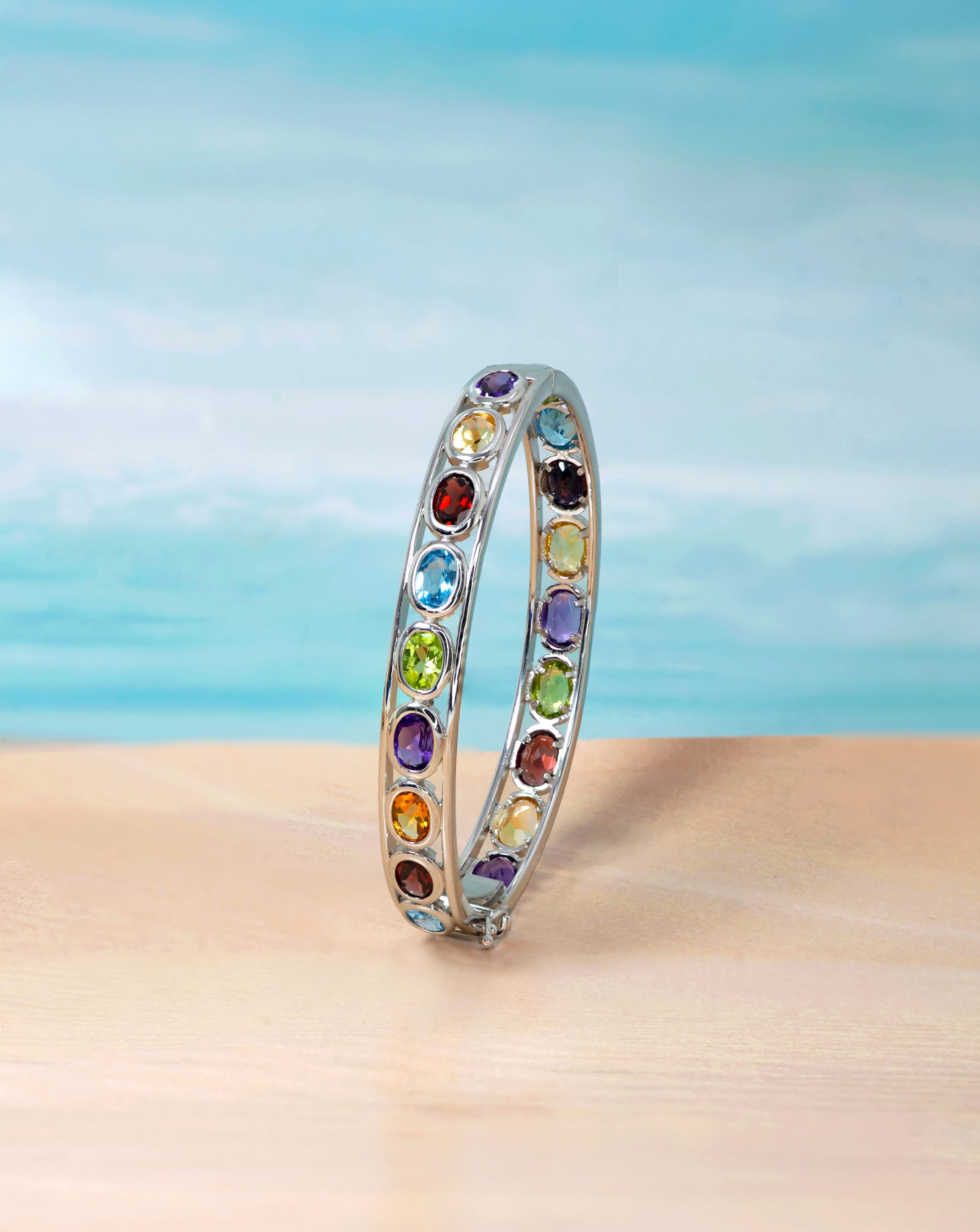 Bangle 3 Curved silver bracelet with rainbow-colored gemstones standing on a sandy beach with a blue ocean backdrop.