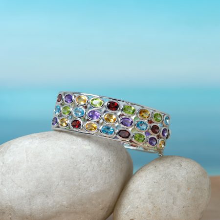 Colorful gem-encrusted bracelet draped over smooth white stones against a blue background.