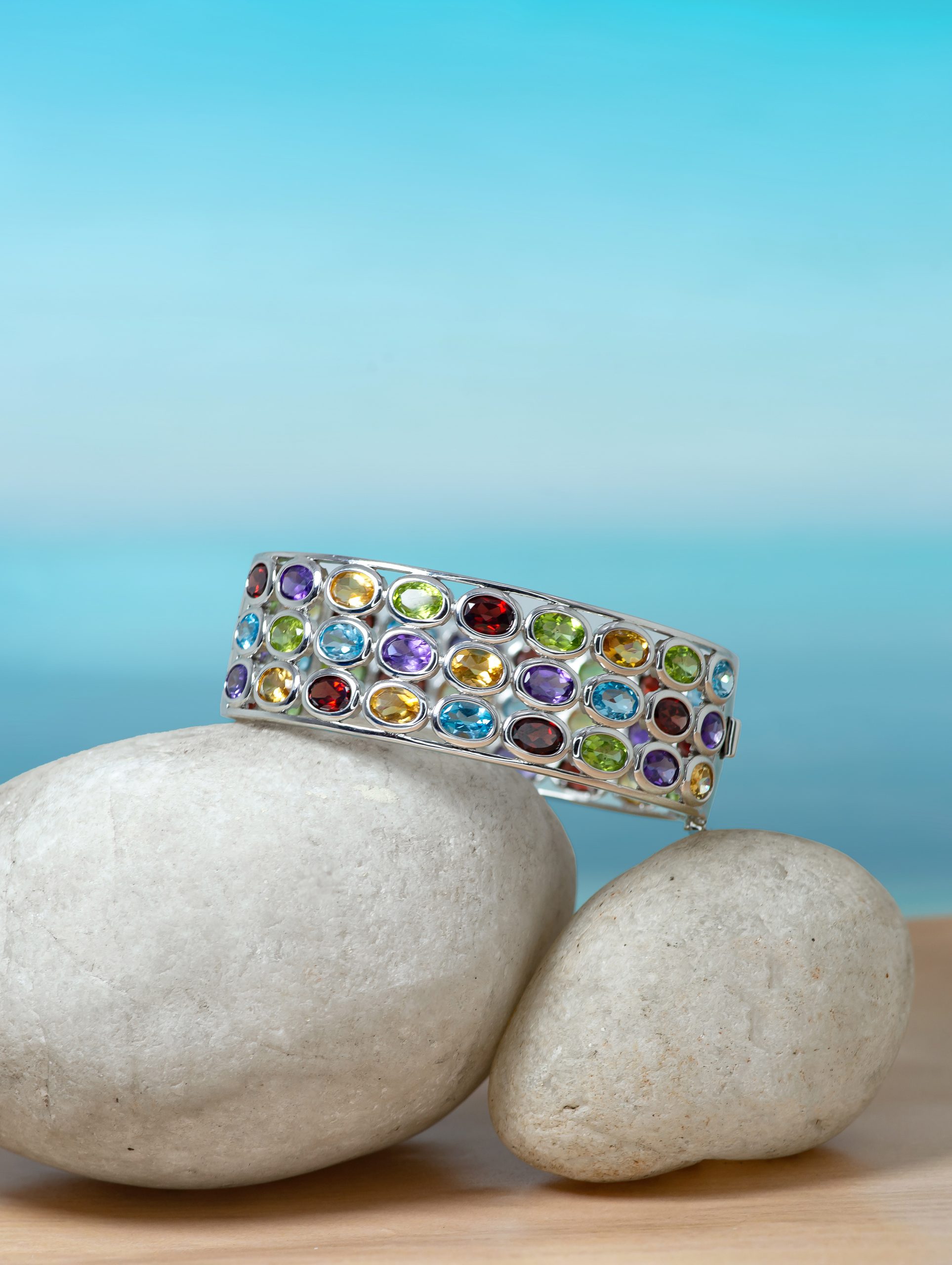 Multi bangle 2 Colorful gem-encrusted bracelet draped over smooth white stones against a blue background.