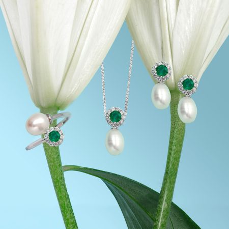Emerald and diamond jewelry hanging from plant stems against a blue gradient background.