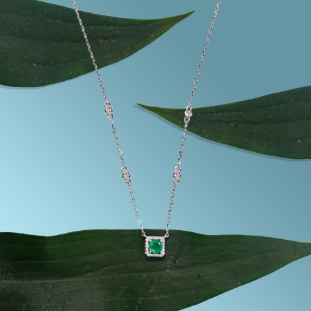 Silver necklace with an emerald-green pendant resting on a broad green leaf against a blue sky backdrop.