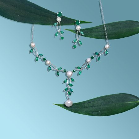 Silver jewelry with emeralds and pearls hanging on green leaves against a blue gradient background (close-up).