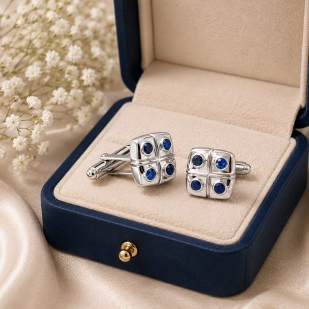 Silver cufflinks with four blue gems in a navy jewelry box, beige lining, beside white flower sprigs.