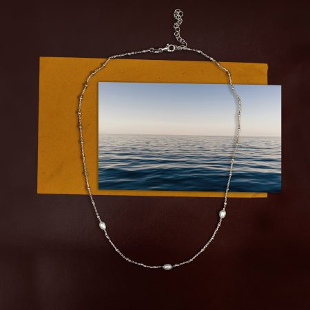 Silver chain necklace with pearl beads laid across a yellow card on a dark brown background, jewelry still life.
