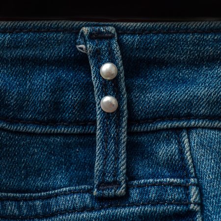 Close-up of blue denim jeans showing a fabric strap with pearl snap buttons and visible stitching.