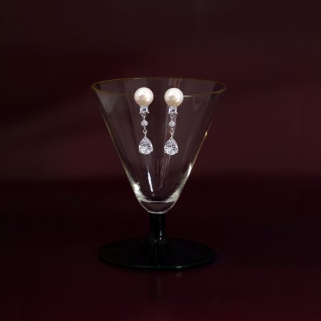 Pearl drop earrings with crystal accents draped over the rim of a wine glass against a dark burgundy background.
