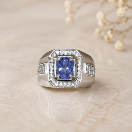 Silver ring with a cushion-cut blue sapphire centerpiece, surrounded by a double halo of diamonds on a beige surface. Inset pavé diamonds decorate the band edges.