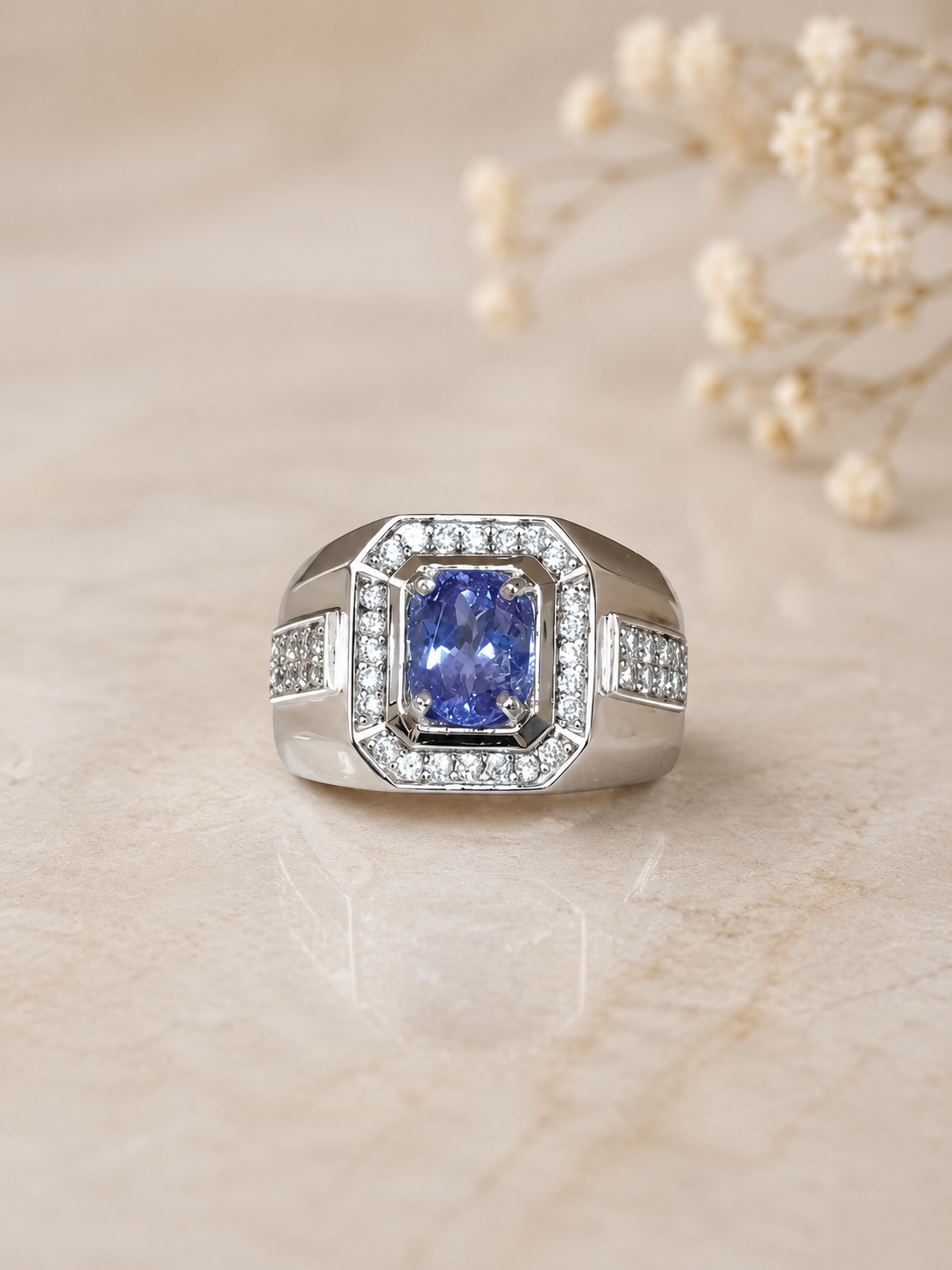 Tanzanite-Ring Silver ring with a cushion-cut blue sapphire centerpiece, surrounded by a double halo of diamonds on a beige surface. Inset pavé diamonds decorate the band edges.