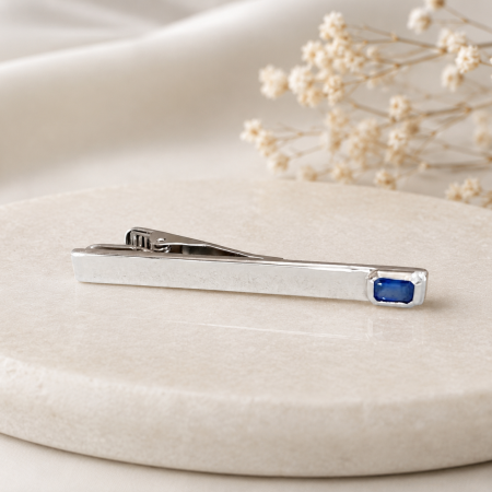 Silver tie clip with a blue gemstone resting on a pale beige display, dried flowers in the background.