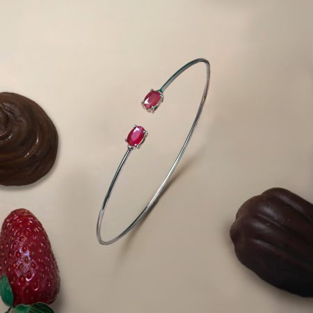 Close-up of a silver bracelet with red gemstone beads resting near strawberries on a pale background, highlighting the jewelry piece.