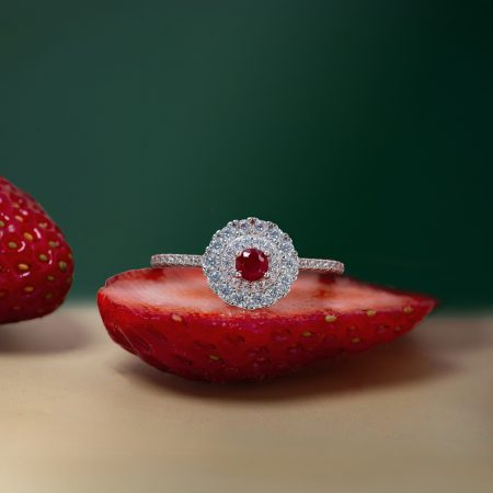 Close-up of a silver ring with a red center stone and surrounding diamonds resting on a halved strawberry against a green background.