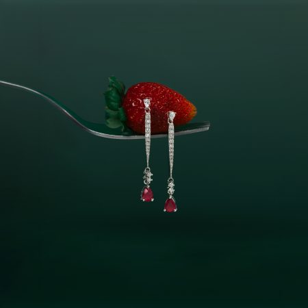 Close-up of a bright red strawberry on a silver fork with dangling diamond jewelry against a dark green background.