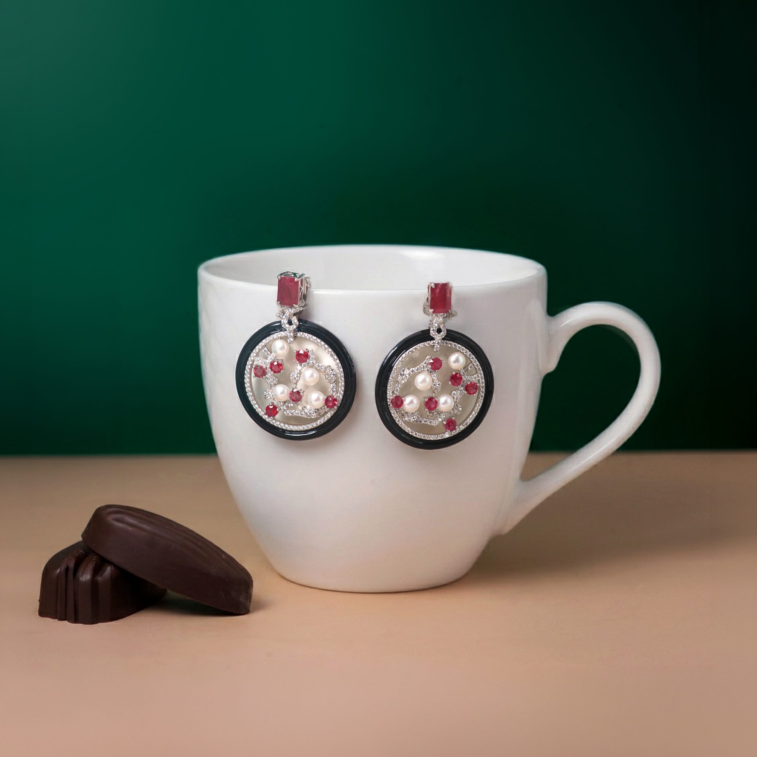 _NZR9754 White mug with dangling jeweled earrings resting on its rim, set against a dark green background with a small chocolate piece on the table nearby.
