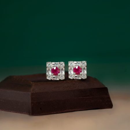 Two small, square-cut diamond earrings with pink center stones set in a silver frame, placed on a dark wooden surface against a green background.