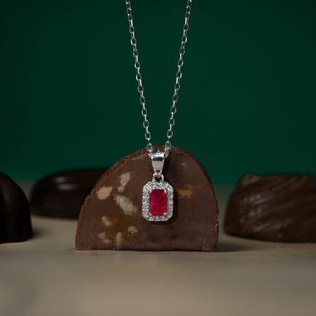 Silver chain necklace with a pink rectangular gemstone surrounded by diamonds, hanging above a chocolate donut backdrop.