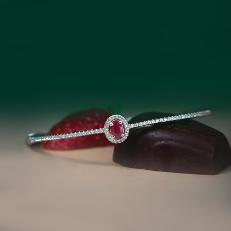 Close-up of a diamond bracelet with a red center stone resting on a brown display surface, green blurred background