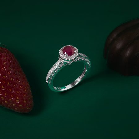 Diamond ring with a red central gemstone surrounded by small diamonds on a green background. A textured red fruit-like object is visible at the edge.