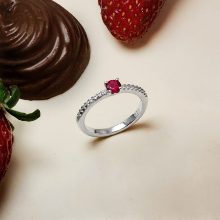 Close-up of a chocolate dessert beside a ripe strawberry and a silver ring with a red gemstone on a pale surface.