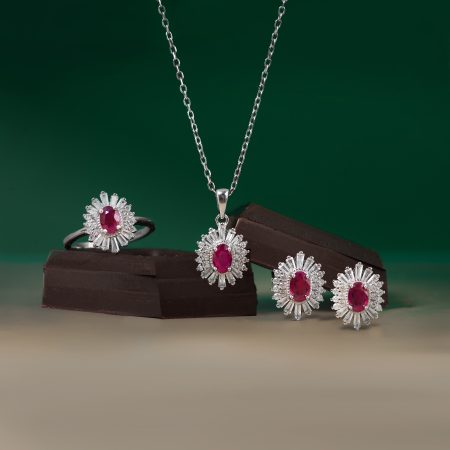 Silver necklace with a ruby-centered pendant surrounded by diamonds, displayed over dark chocolate bars against a green background and jewelry nearby.