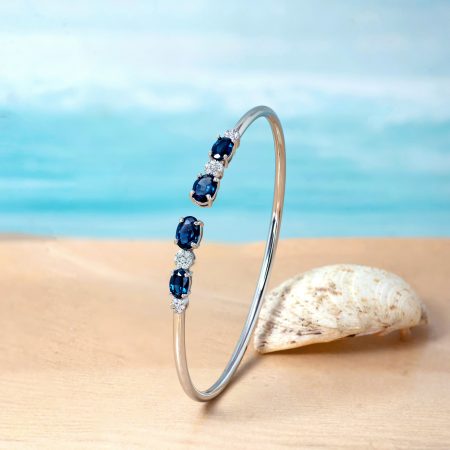 Delicate silver bracelet with dark blue gemstones and small diamonds displayed on a curved stand by a seashell on the sandy beach
