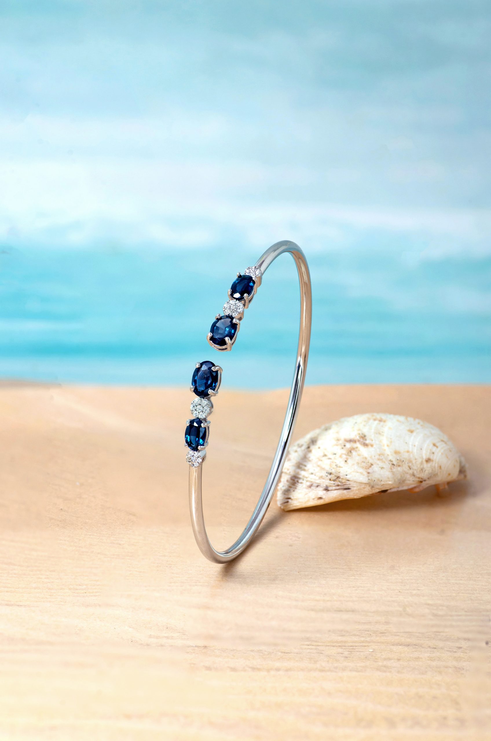 Bangle 1 Delicate silver bracelet with dark blue gemstones and small diamonds displayed on a curved stand by a seashell on the sandy beach