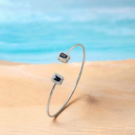 Two silver rings or bangles with square black stones surrounded by diamonds displayed outdoors on a sandy beach.