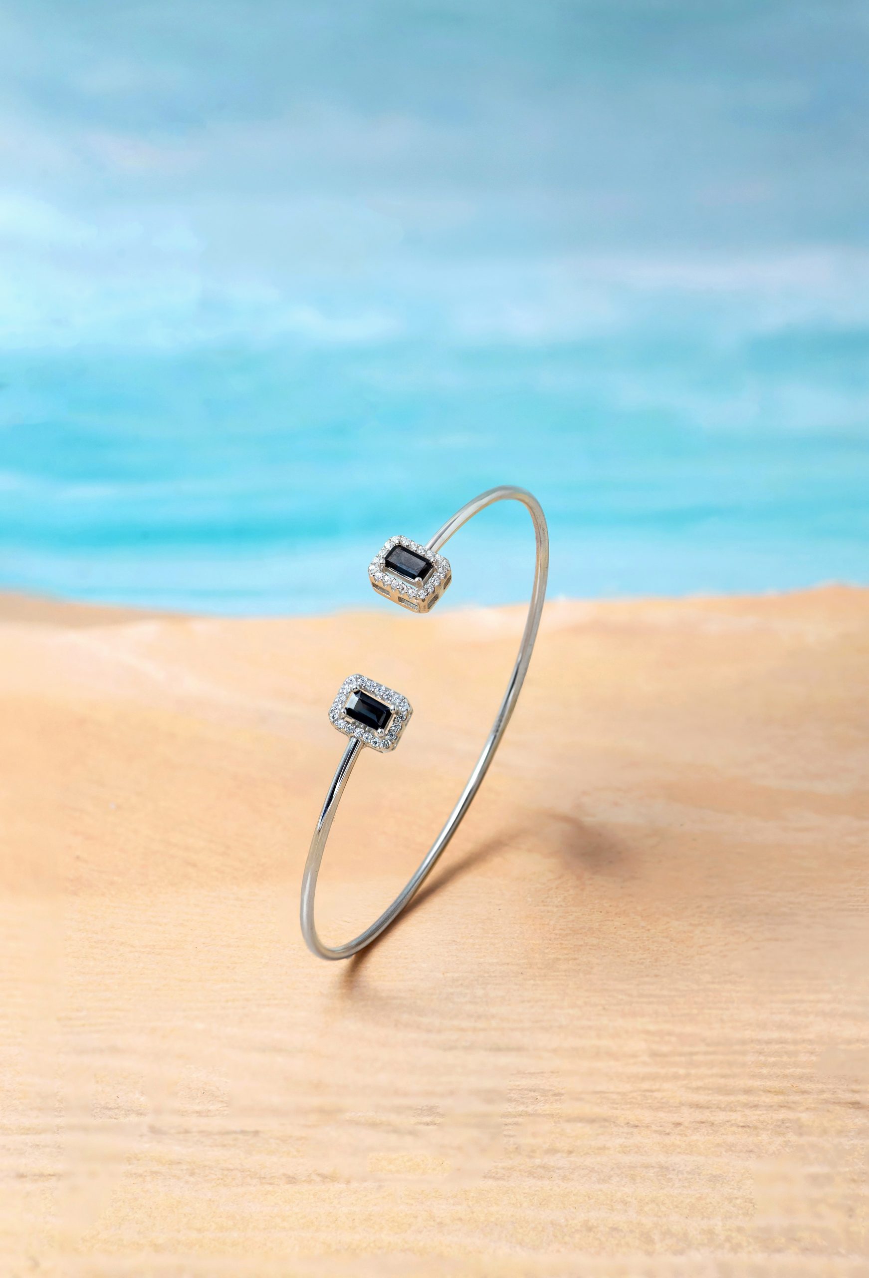 Bangle 2 Two silver rings or bangles with square black stones surrounded by diamonds displayed outdoors on a sandy beach.