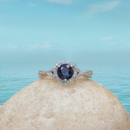 Diamond ring with a dark blue center stone set in a halo of small diamonds, resting on a smooth beige rock with a turquoise sea and sky backdrop