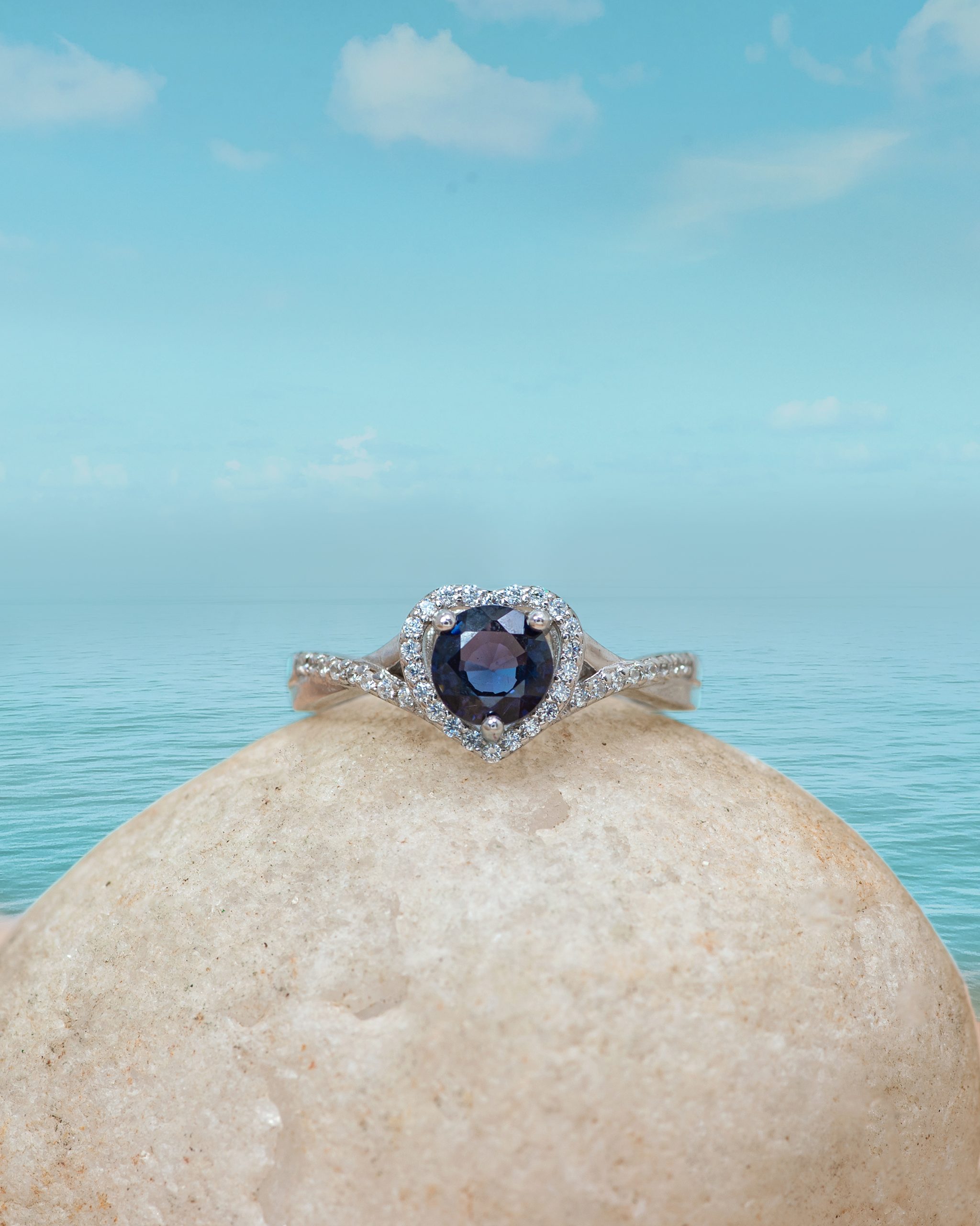 Heart Ring Diamond ring with a dark blue center stone set in a halo of small diamonds, resting on a smooth beige rock with a turquoise sea and sky backdrop