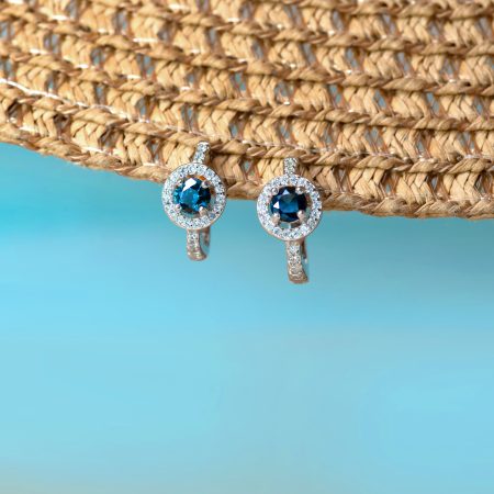 Pair of silver earrings with round blue stones surrounded by a halo of small diamonds, hanging against a woven straw background and blue gradient.