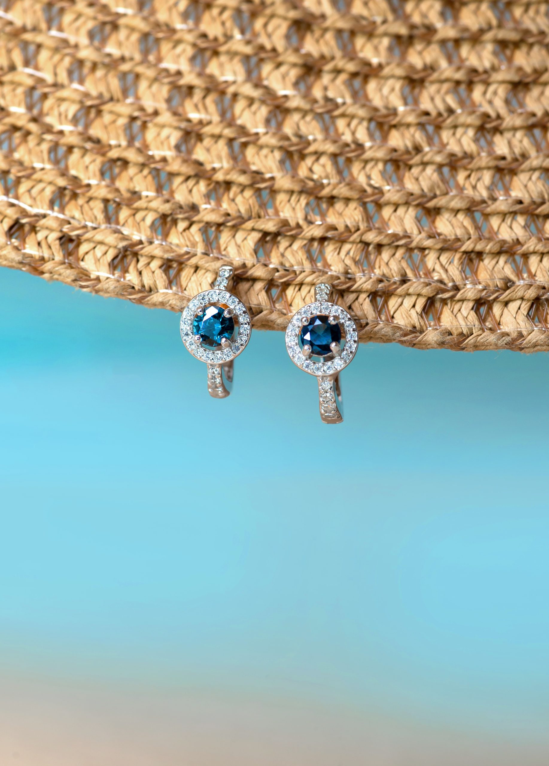 _NZR0276 Pair of silver earrings with round blue stones surrounded by a halo of small diamonds, hanging against a woven straw background and blue gradient.