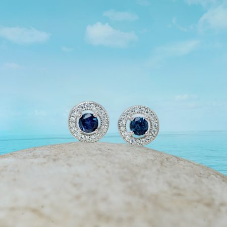 Two halo-style silver rings with deep blue center stones, set on a sandy rock by the sea under a blue sky.