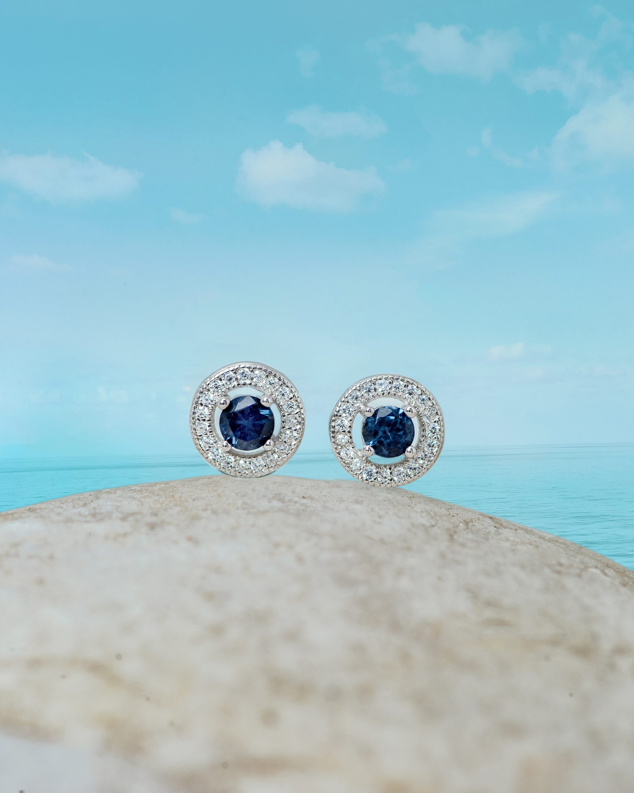 _NZR0535 Two halo-style silver rings with deep blue center stones, set on a sandy rock by the sea under a blue sky.