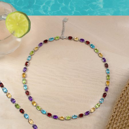 Long necklace with multicolored gemstones laid in a loose arc on a beige surface, with a lime drink and pool in the background? (informative)