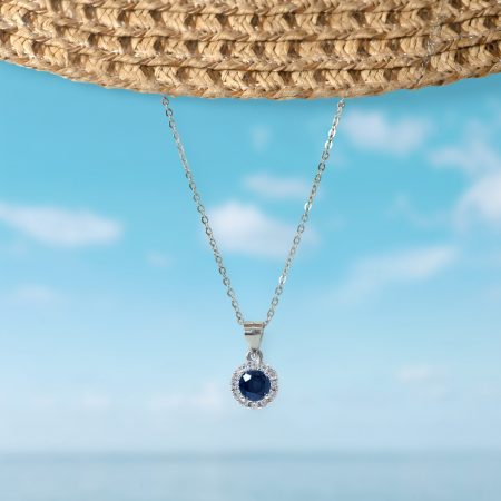 Silver necklace with a dark blue gemstone pendant surrounded by small diamonds, hanging against a blue sky.
