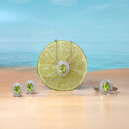 Close-up of lime slice jewelry: a green gemstone pendant with diamond halo on a silver chain resting on a sandy beach, with additional matching green-stone rings nearby.