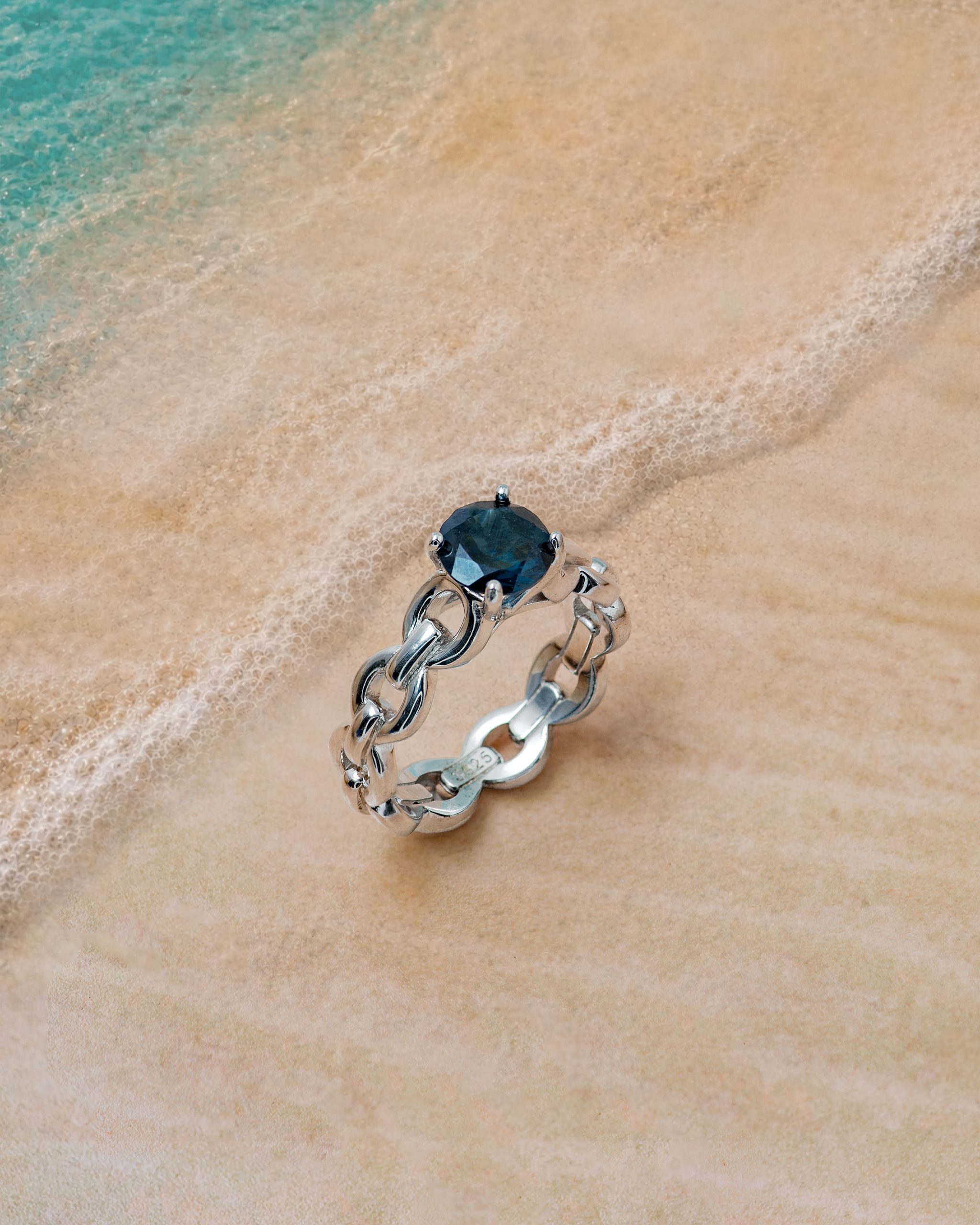 Ring 2 Silver bracelet with a dark blue gemstone resting on wet sand by the ocean.
