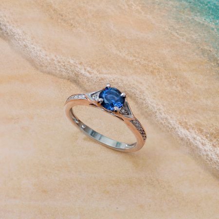 Close-up of a silver ring with a blue gemstone resting on wet sandy beach with turquoise water in the background.
