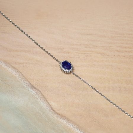 Silver chain necklace with a deep blue oval gemstone pendant surrounded by small diamonds on a beige fabric background