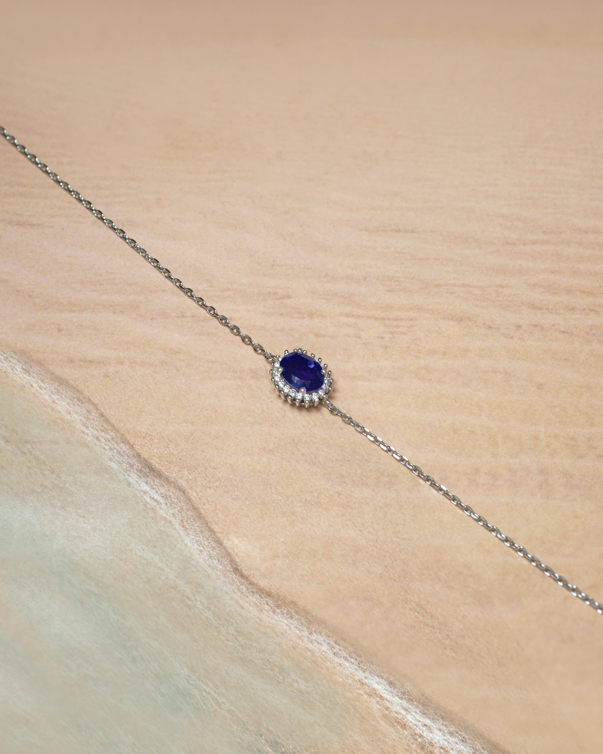 bracelet Silver chain necklace with a deep blue oval gemstone pendant surrounded by small diamonds on a beige fabric background