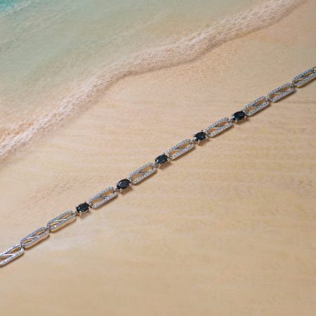 Silver necklace with dark blue stones laid along a sandy beach by turquoise water. (informative, describes function and scene)