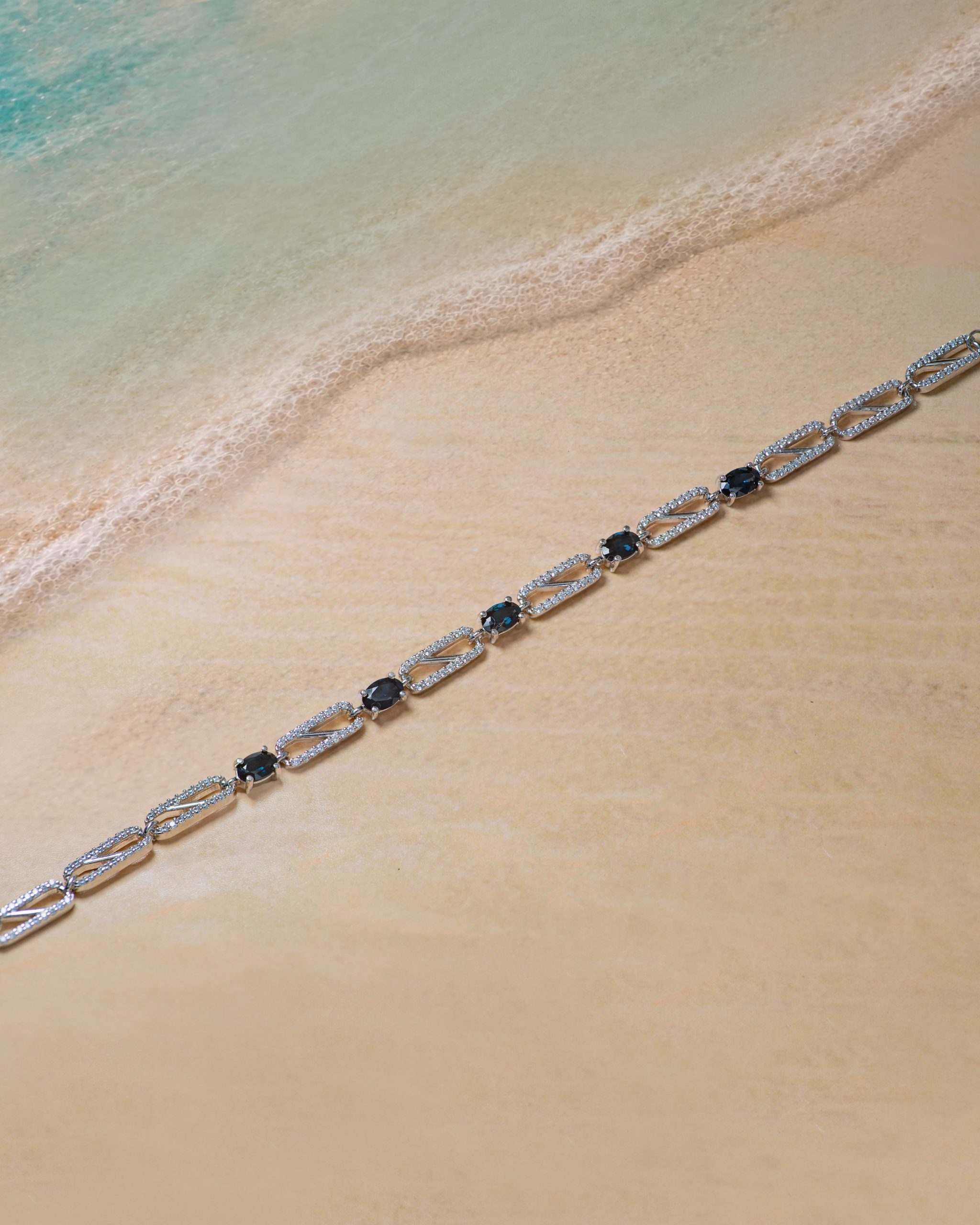 braceletb Silver necklace with dark blue stones laid along a sandy beach by turquoise water. (informative, describes function and scene)